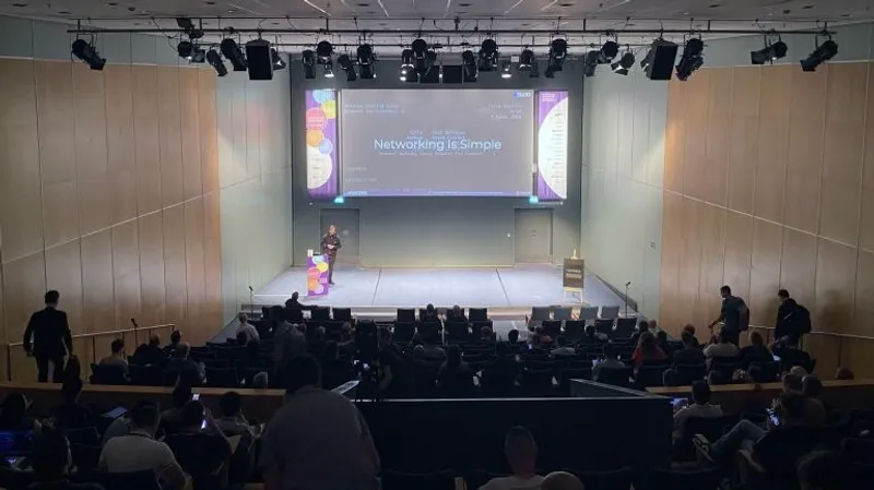 Conference presentation showing "Networking is Simple" slide in modern auditorium with Vlad Iliushin from ELLIO as a speaker on stage and audience seated in tiered seating