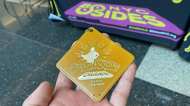 Hand holding a golden square BSides NYC 2025 badge with TRAINER designation