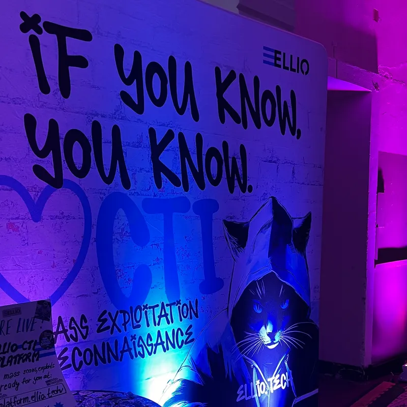Cybersecurity-themed wall mural with "IF YOU KNOW, YOU KNOW" text and cat figure in hoodie, displaying "MASS EXPLOITATION RECONNAISSANCE" and ELLIO branding in purple/blue lighting