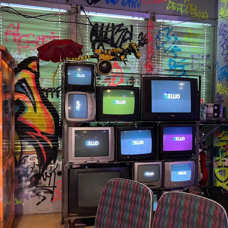Stack of vintage CRT televisions displaying ELLIO logos in a graffiti-covered room with colorful street art on walls and security shutters