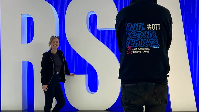 Two ELLIO founders Jana Tom and Vlad Iliushin at RSA conference, one wearing ELLIO hoodie with "HUNT. RESPOND. ELIMINATE" and ELLIO logo, standing by large "RSA" letters